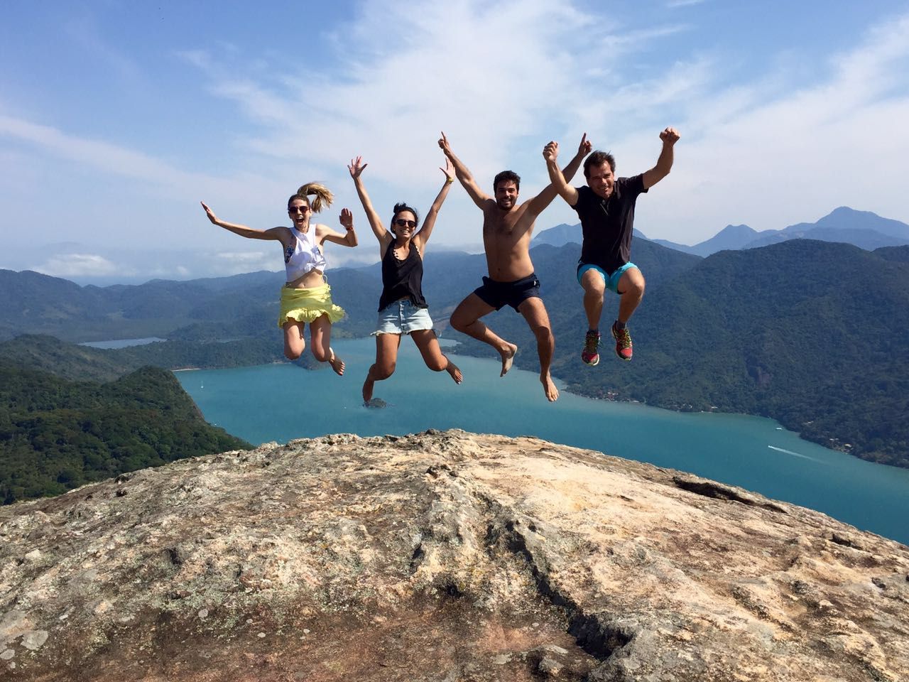 Cuatro amigos saltando en la cima del Pão de Açúcar do Mamanguá, con el brazo de mar y las montañas al fondo