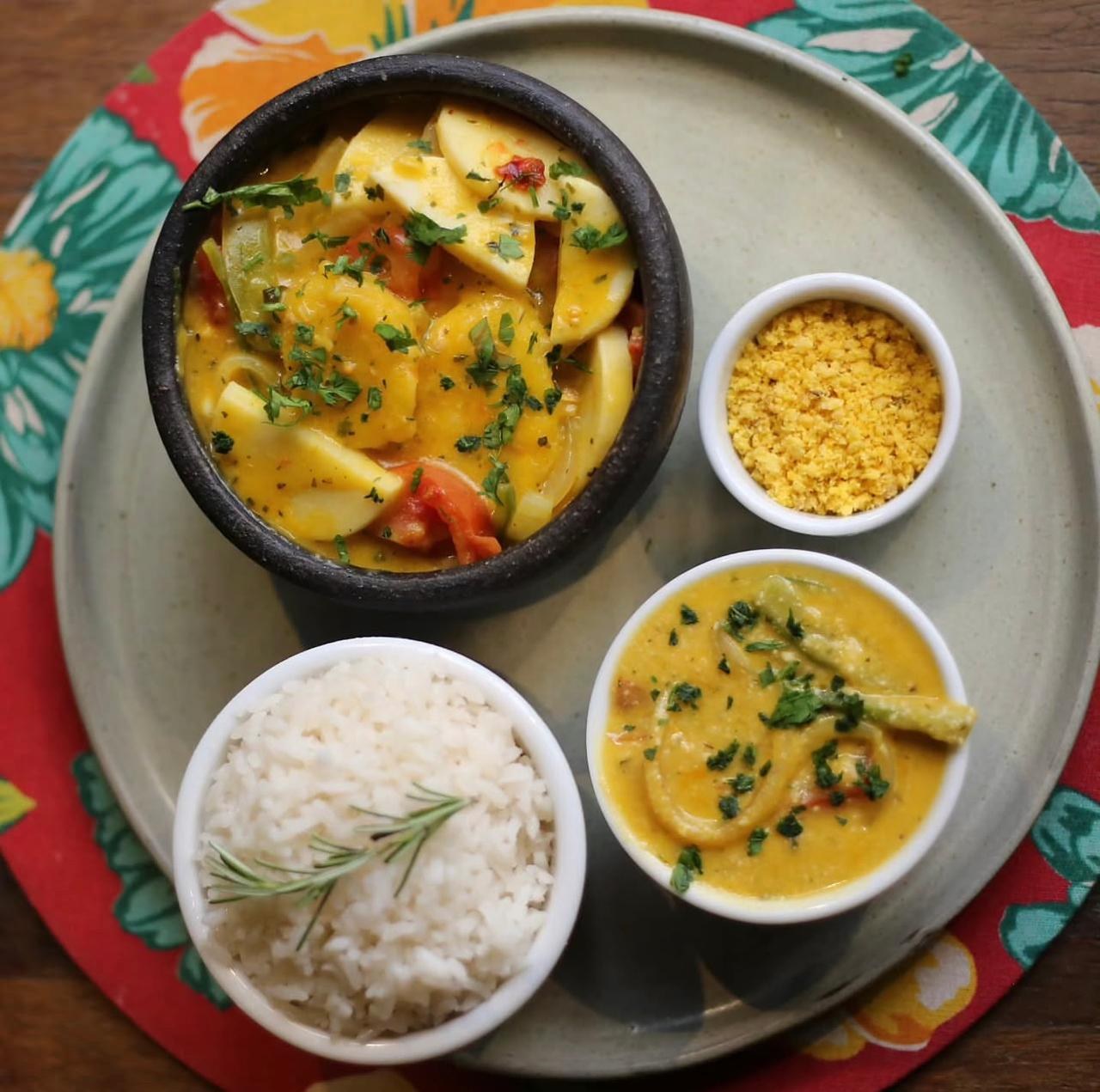 Moqueca vegana en una olla de piedra: rodajas de plátano macho y palmito en caldo amarillo de dendê, con arroz, farofa y pirão al lado