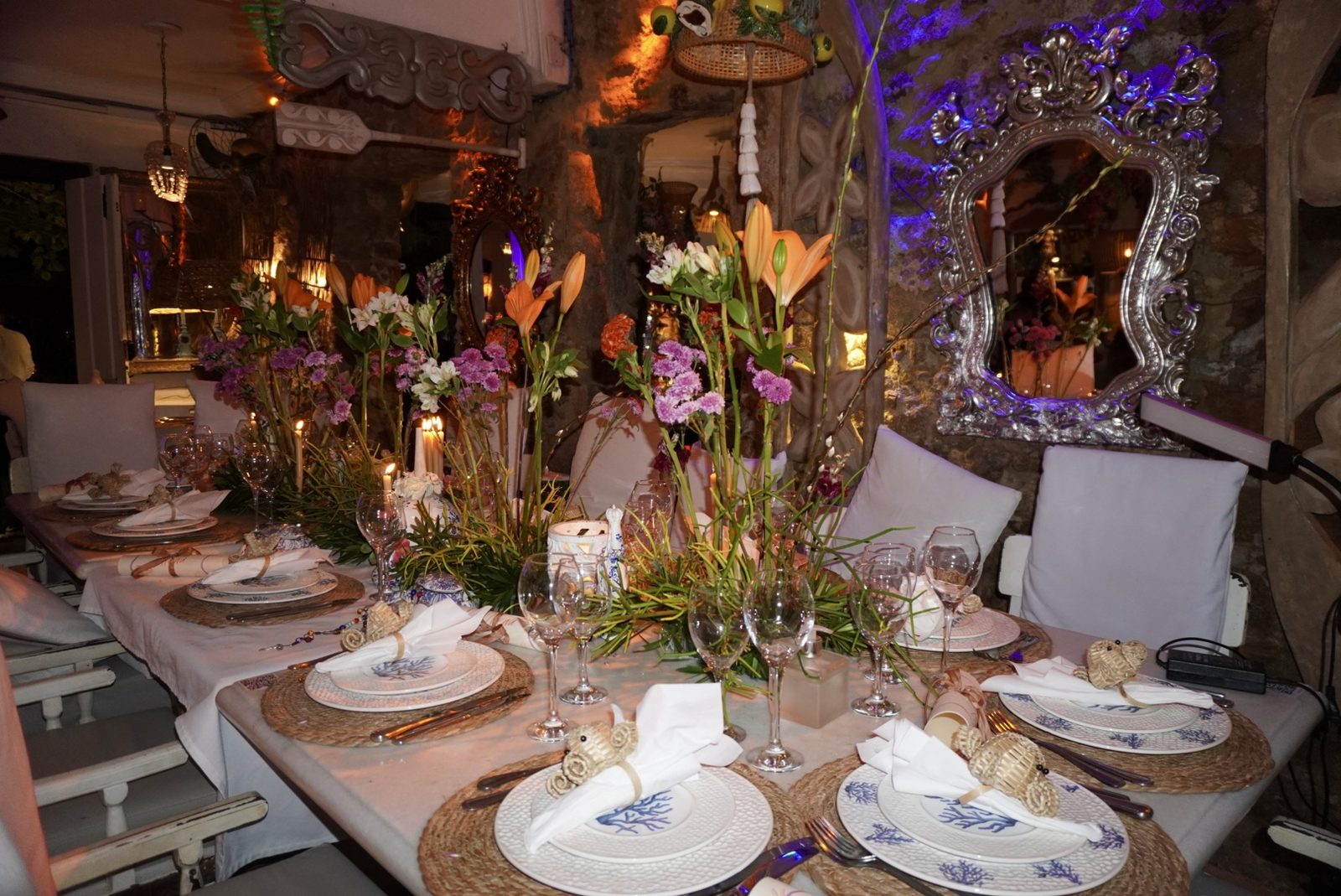 Table dressée au Refúgio : assiettes bleu corail sur sets en raphia, fleurs, bougies et un miroir ancien sur le mur de pierre