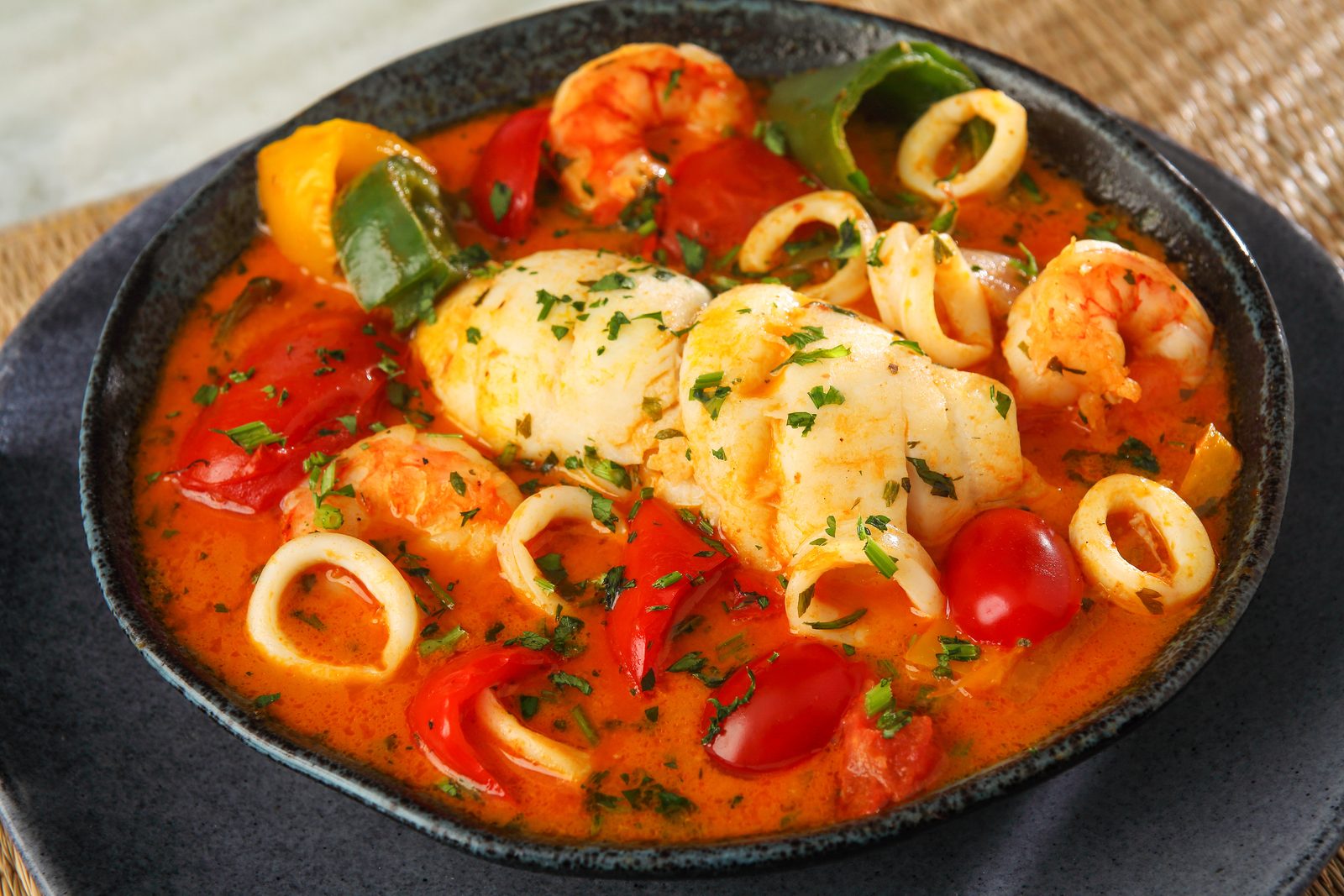 Moqueca de fruits de mer dans une cocotte sombre : poisson blanc, crevettes, anneaux de calamar, poivrons et tomate